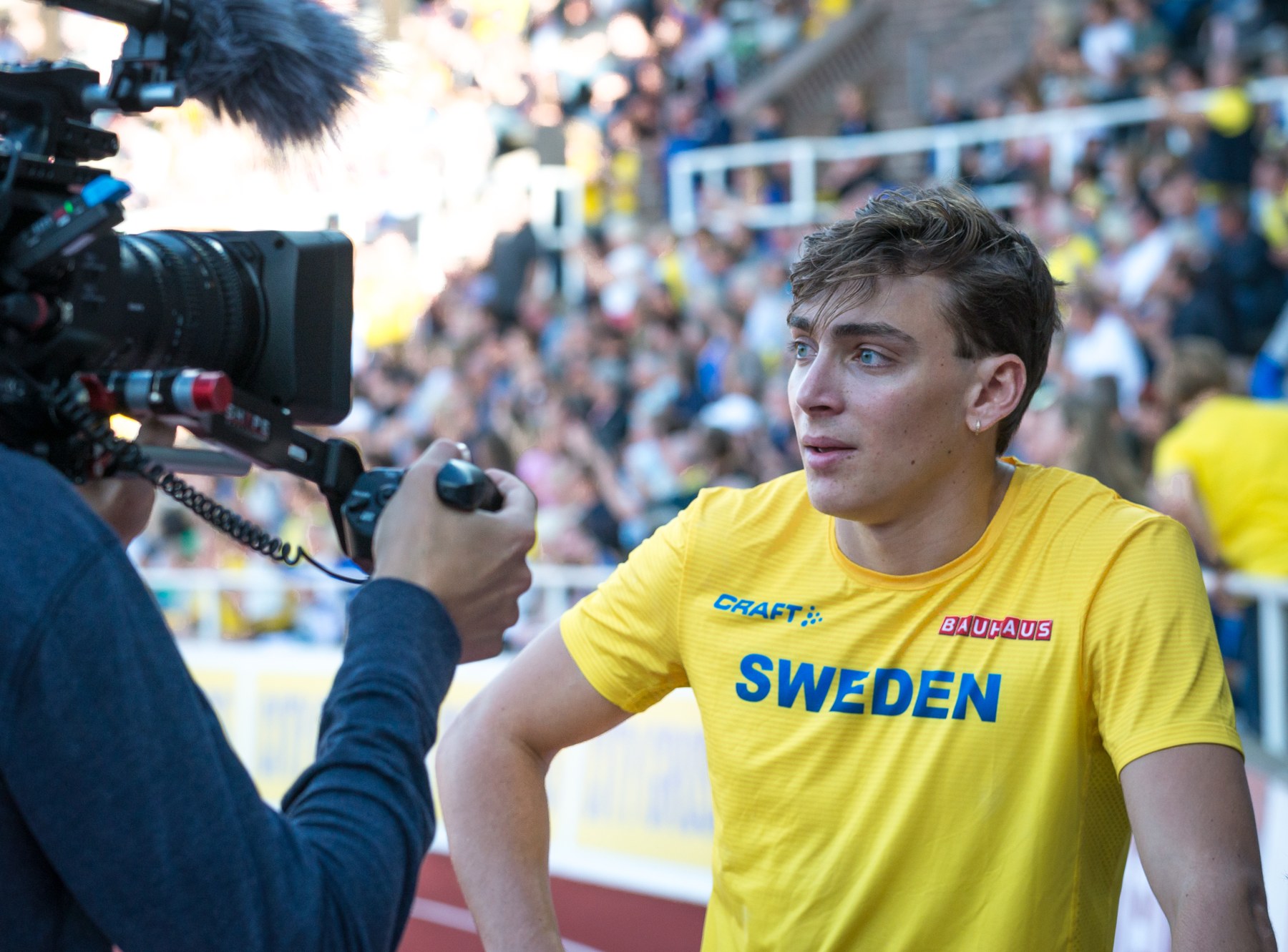 Armand Duplantis in a yellow Sweden shirt speaking beside a television camera.
