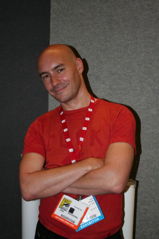 Grant Morrison in a red shirt and convention badge, with arms folded.