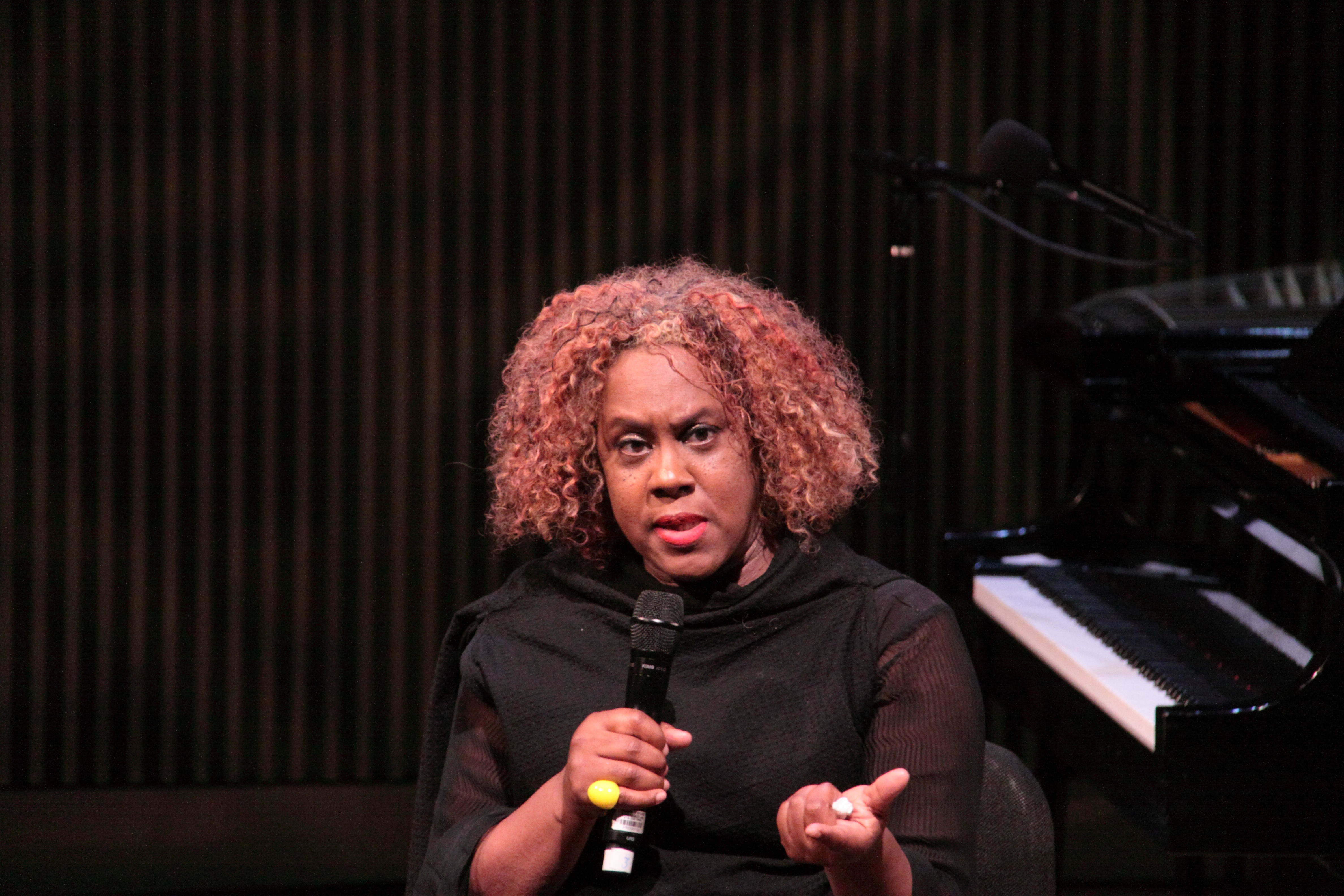 Errollyn Wallen holds a microphone beside a piano on a dark stage.