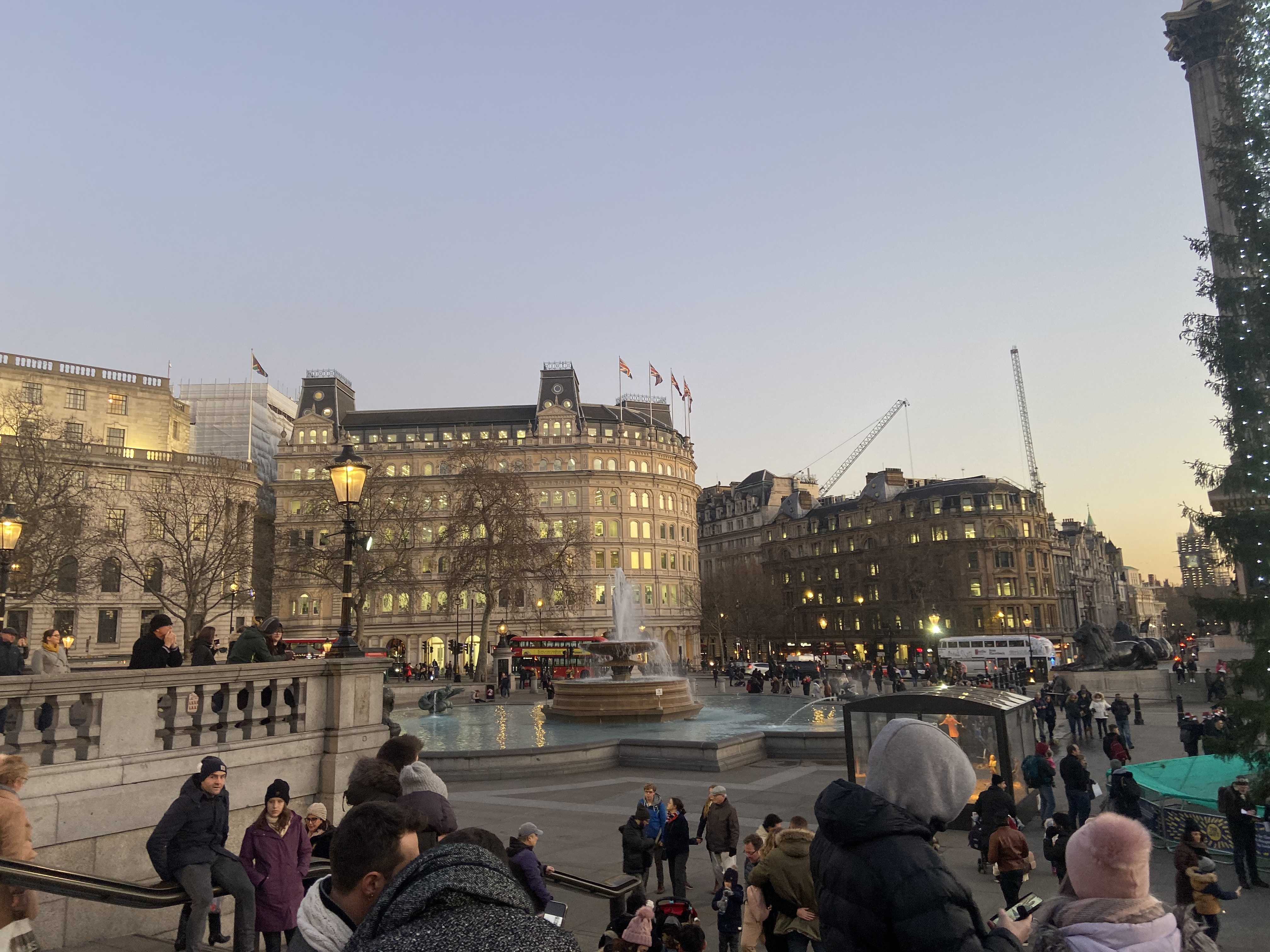 Visitors stand near a lit fountain and buildings at dusk.