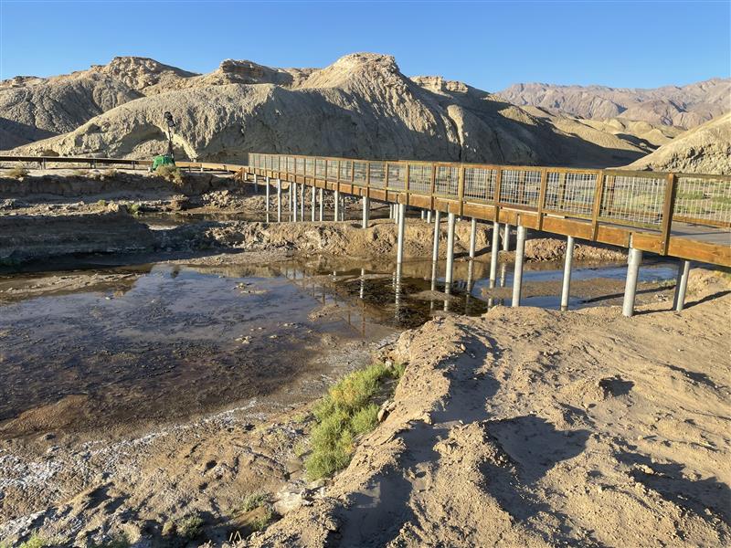 A wooden boardwalk on pale supports spans a shallow desert creek beneath barren hills.