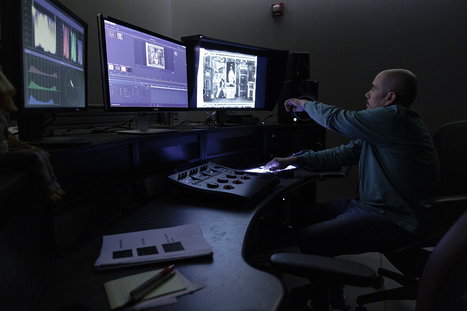 A film-restoration worker sits at grading controls and monitors showing a black-and-white film.