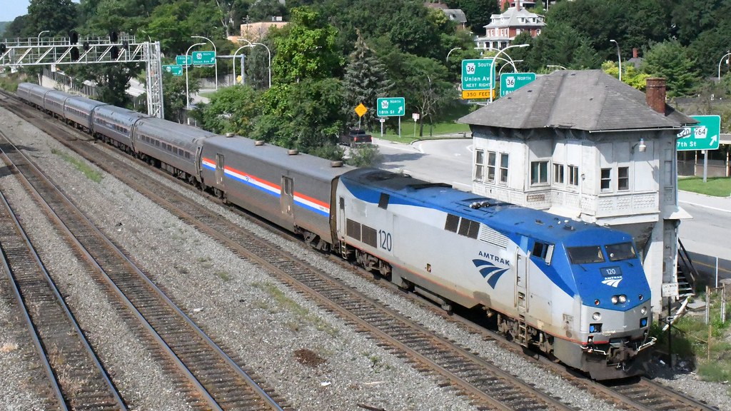 An Amtrak locomotive pulls passenger cars past a railway building.