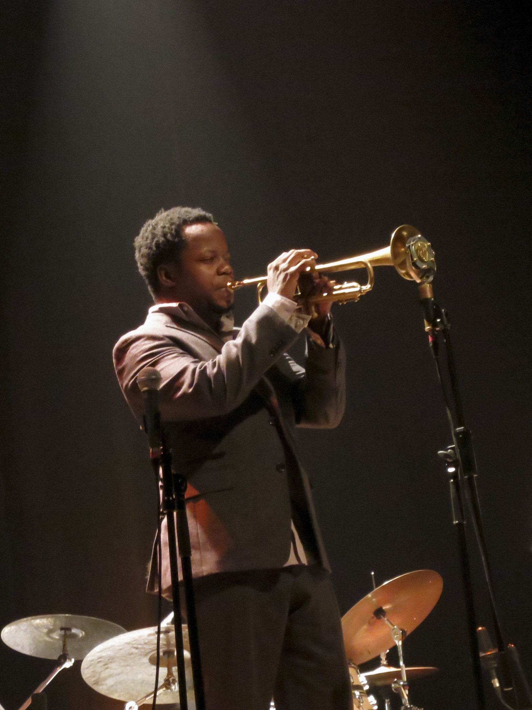 Ambrose Akinmusire plays a trumpet onstage in a gray jacket, with drums behind him.