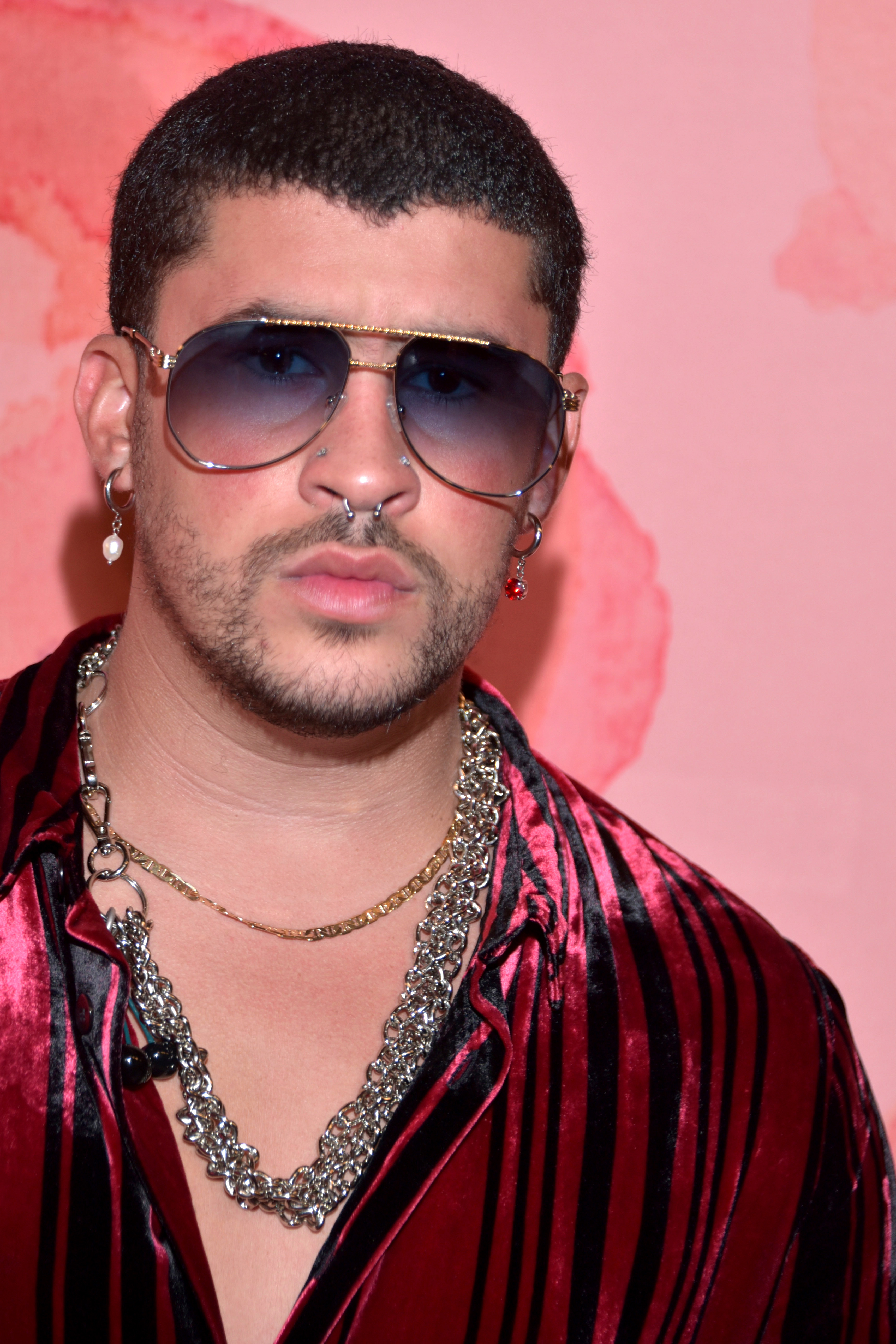 Bad Bunny wears sunglasses, layered chains and a red-and-black striped shirt in a file portrait.