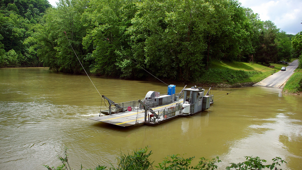 A cable-guided vehicle ferry approaches a wooded riverbank.