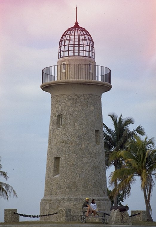 Boca Chita Key’s stone lighthouse beside palms, with two visitors near its base.
