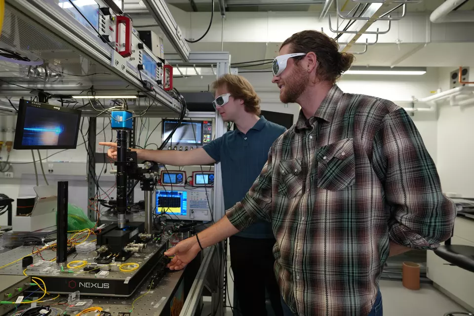 Two researchers in protective eyewear work beside optical equipment and instrument displays.