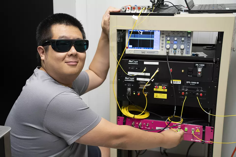 A researcher in protective glasses stands beside a rack of photon-distribution equipment.