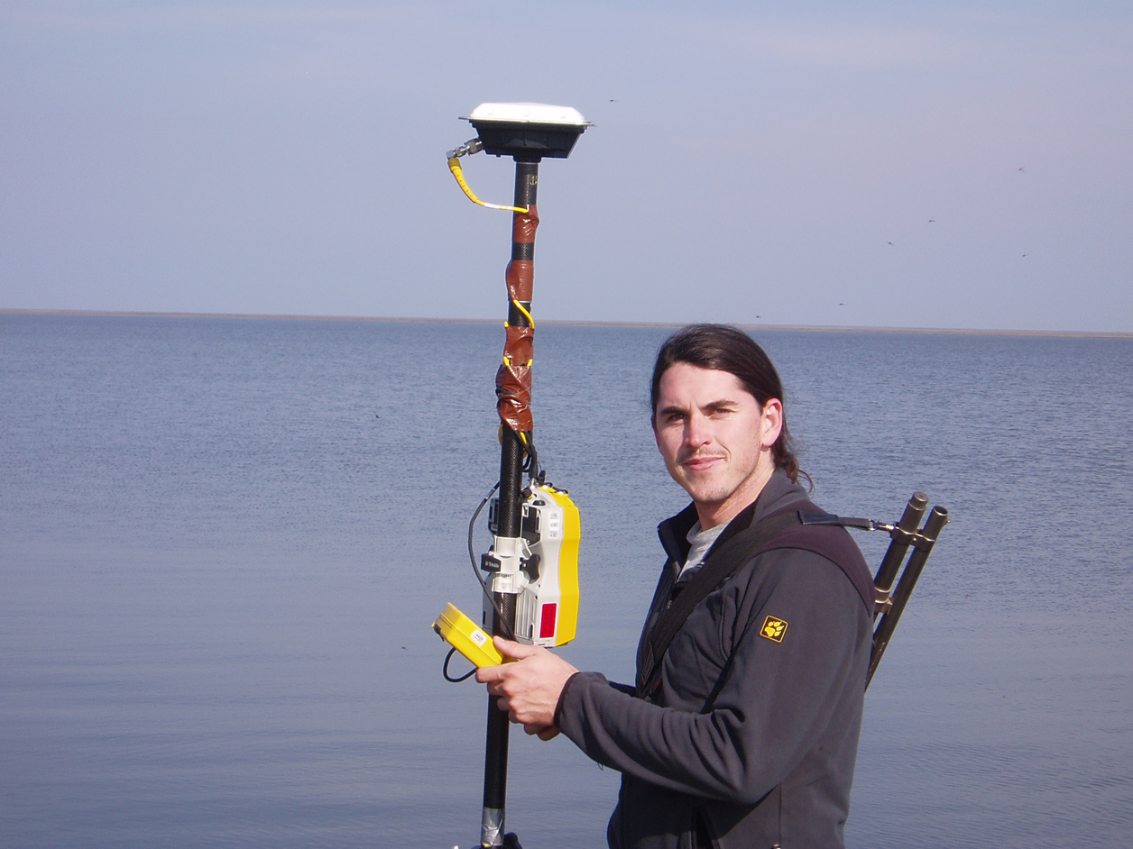 Man holding a GPS survey pole beside water.