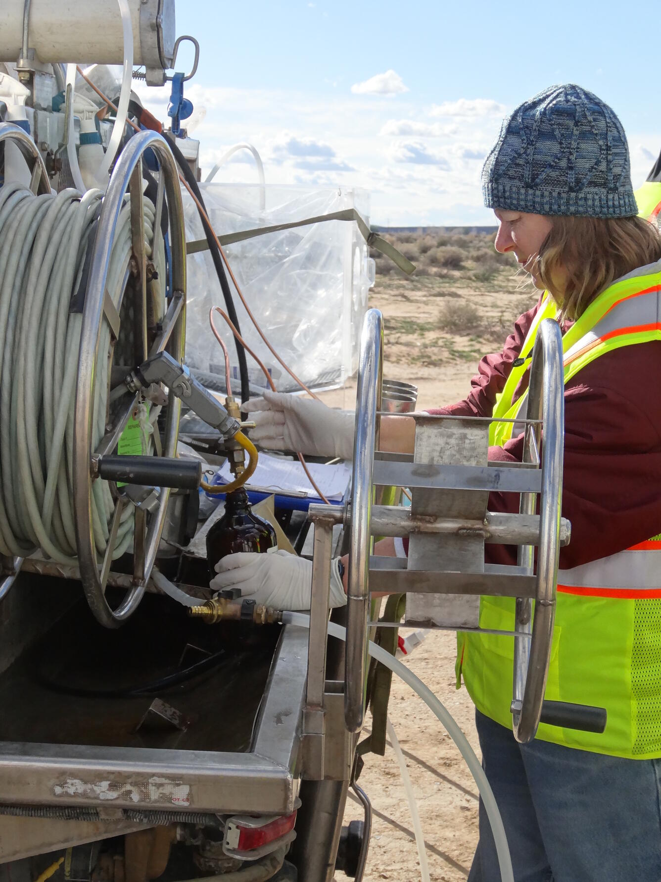 A gloved hydrologist collects water in a sample bottle beside well-sampling equipment.