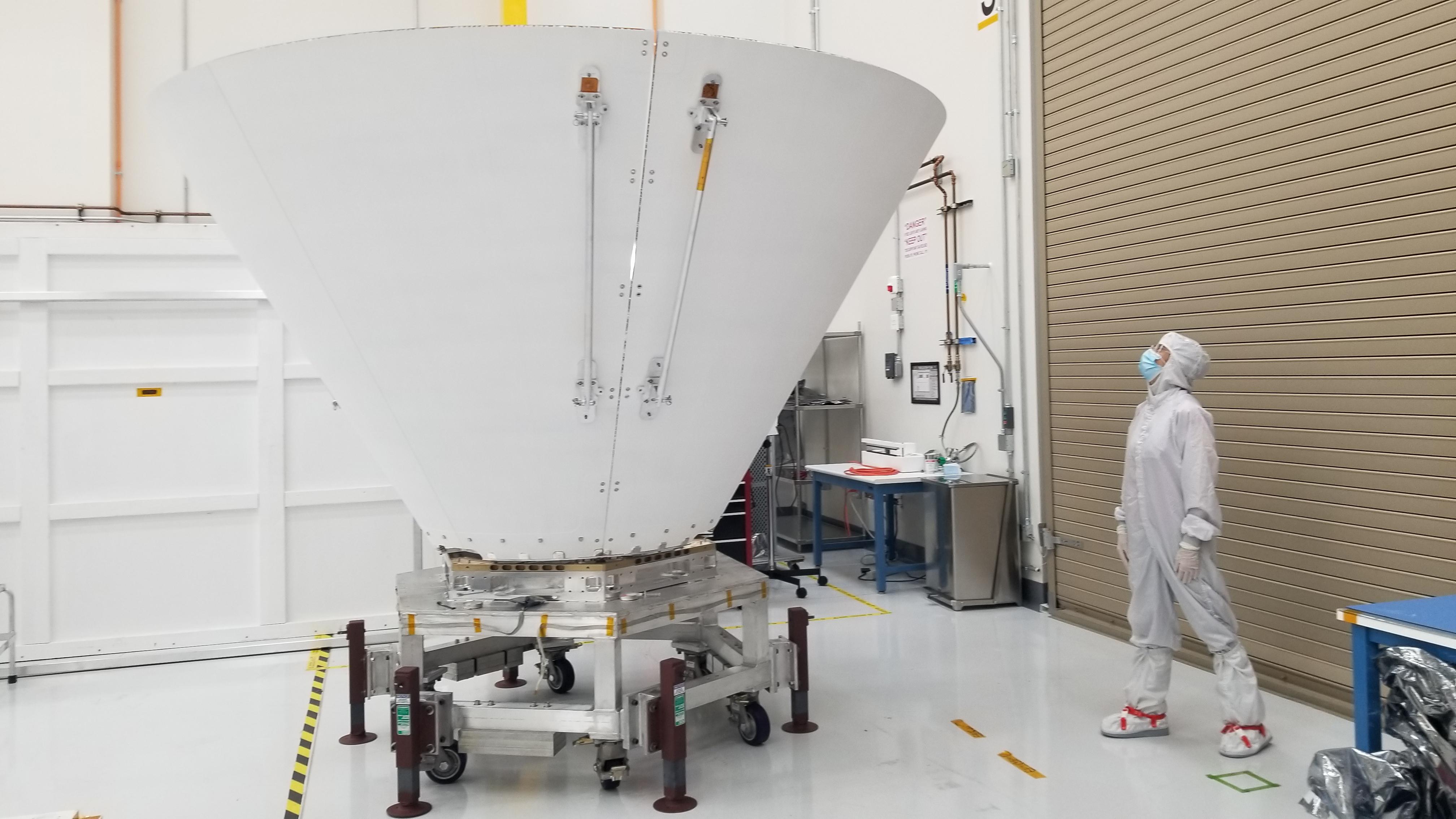 A large conical spacecraft shield stands beside a cleanroom worker.