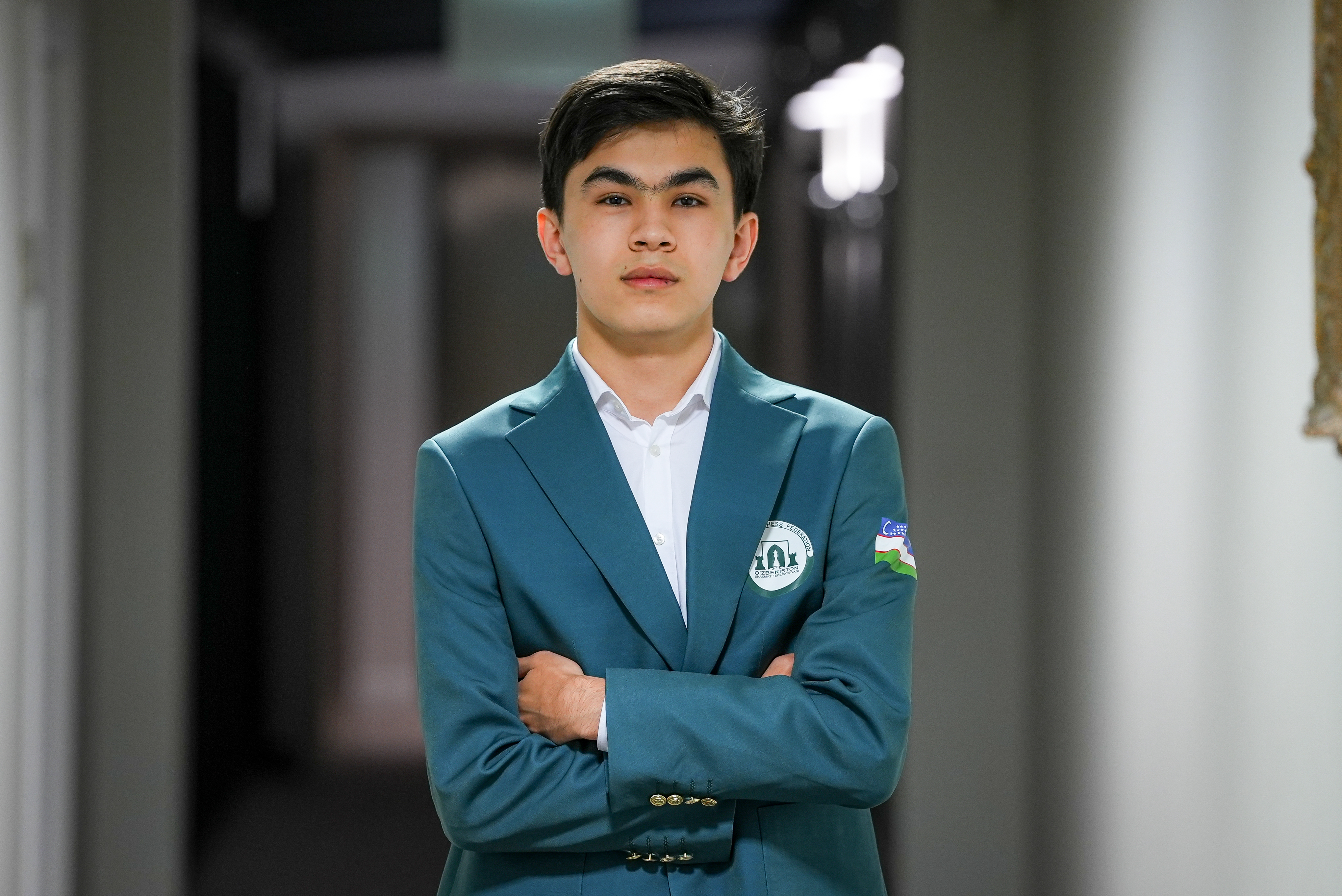 Sindarov wears a teal Uzbekistan chess federation blazer with his arms crossed.