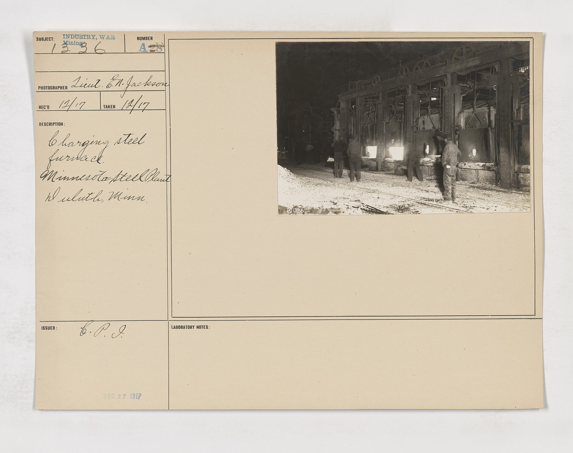 Archival photograph mounted on a labeled card showing workers charging a steel furnace.