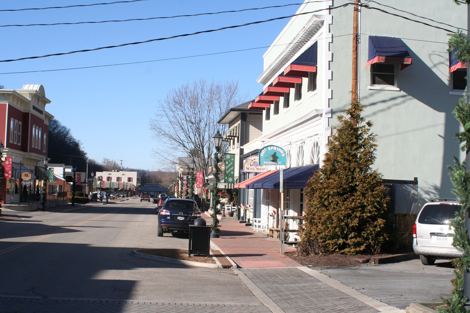 A downtown street with brick and painted storefronts, parked cars and overhead wires.