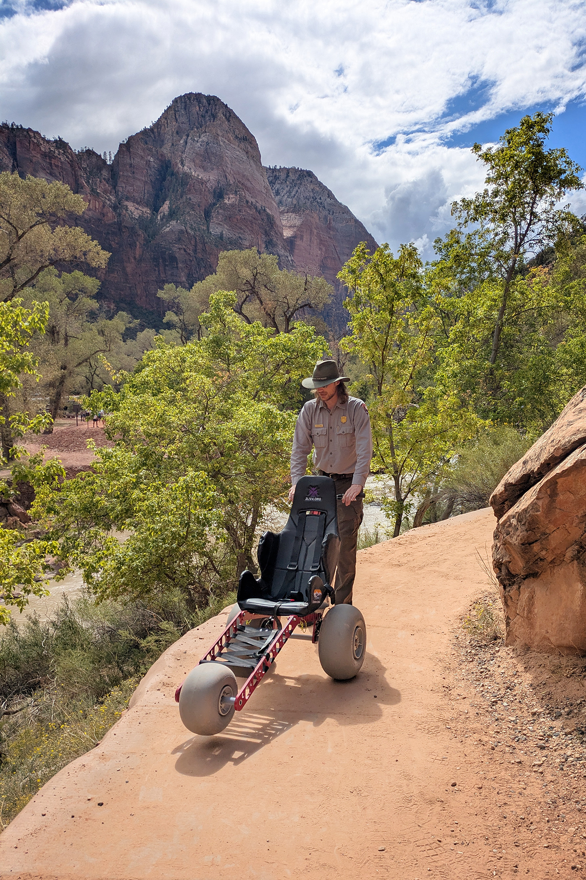 Ranger stands behind an empty three-wheeled chair on a trail.