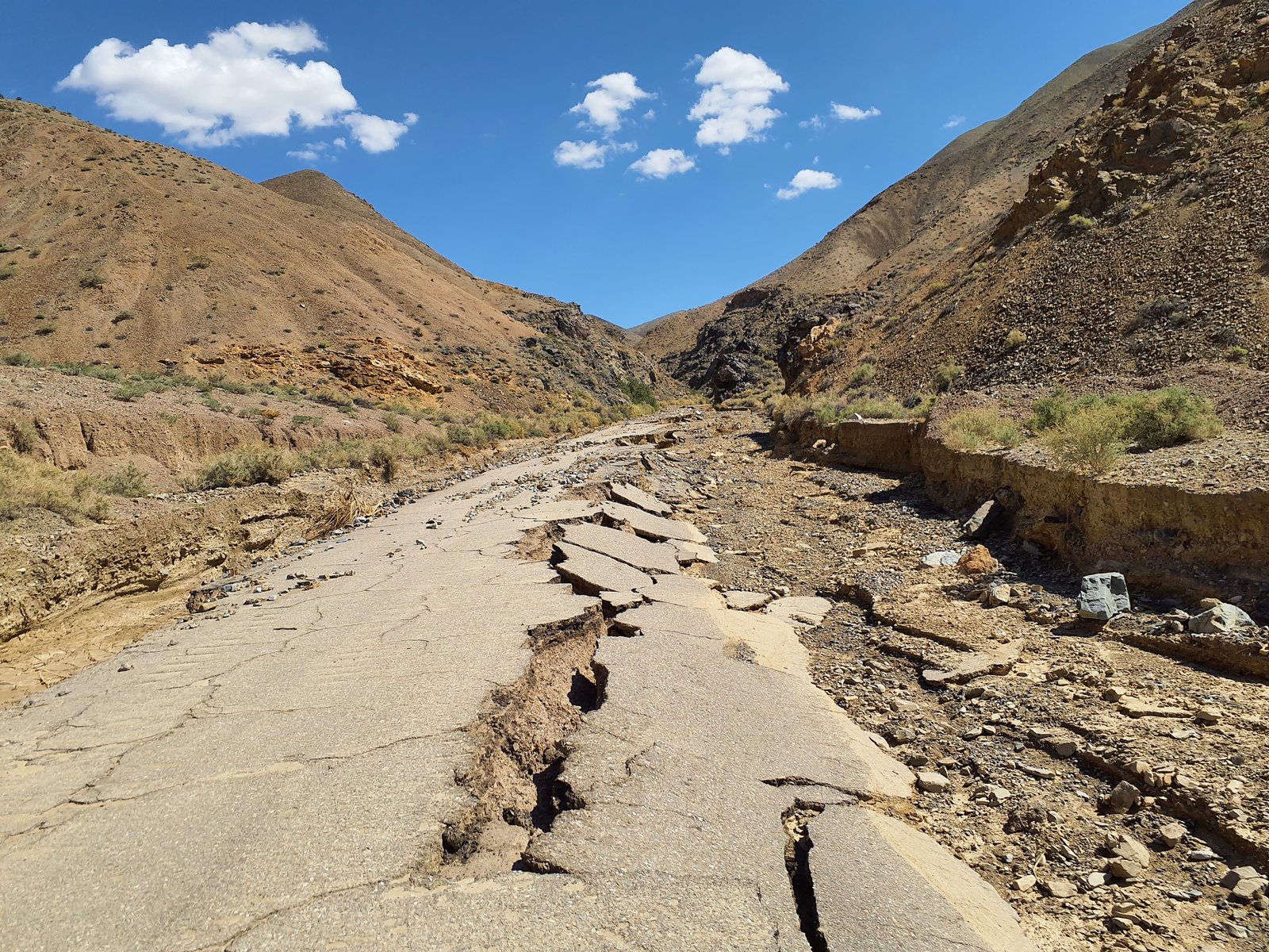 Broken asphalt and deep erosion cuts run along a desert road between rocky hills.